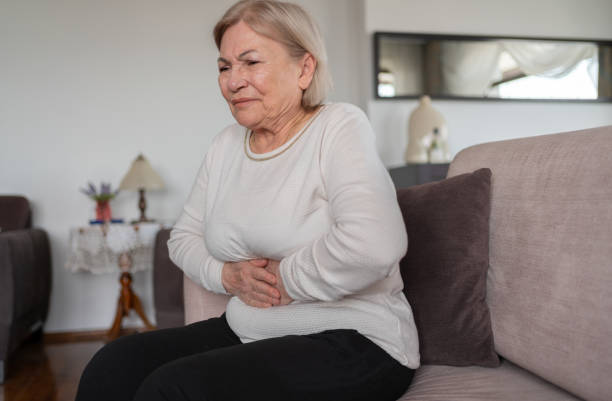 An older woman sits on a couch, visibly distressed, holding her stomach in pain, conveying a sense of discomfort and concern.