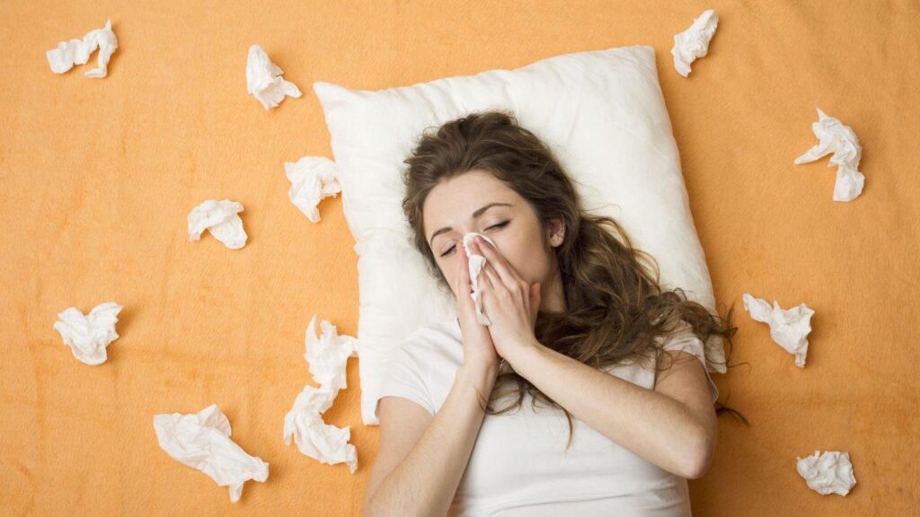 A sick woman lays on a bed and blows her nose into a tissue.