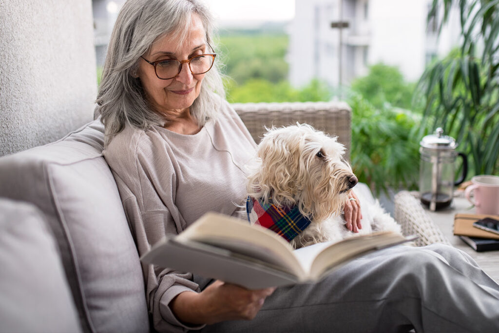 An older woman with glasses is reading a book while her dog sits beside her, creating a cozy and serene atmosphere.