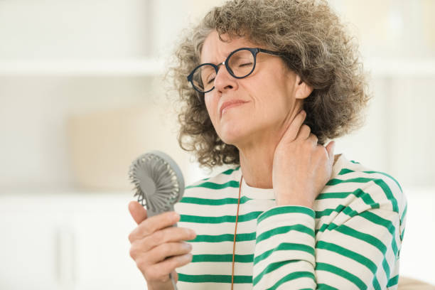 A woman with curly hair wears glasses and holds a handheld fan while resting her hand on her neck, dressed in a striped shirt.