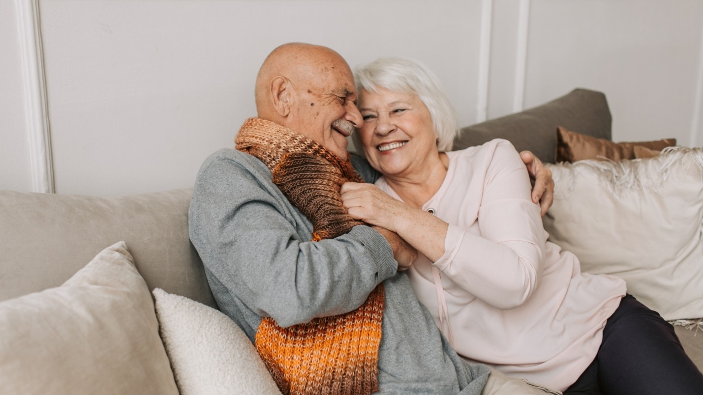 An elderly couple smile and sit on a couch together.